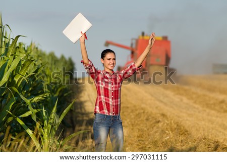 Cheerful beautiful girl in plaid shirt standing in wheat field with raised arms, combine harvester in background