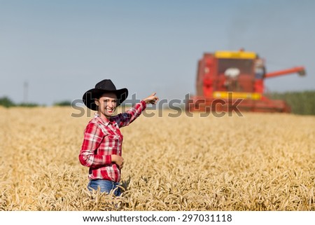 Pretty cowgirl with hat standing in field and showing combine harvester working in wheat field