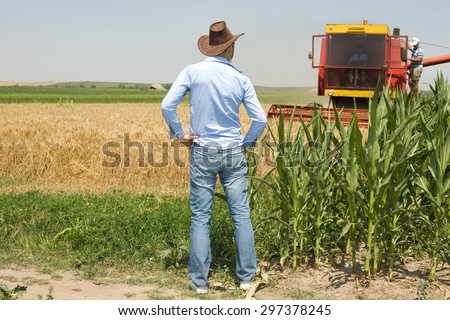 Young attractive farmer with cowboy hat standing in the field and looking at combine harvester