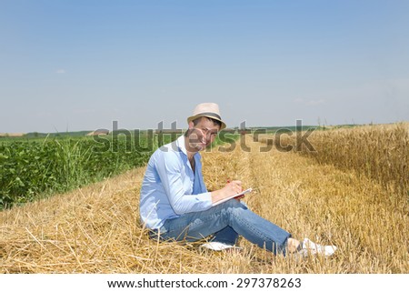 Young attractive man sitting on haystack on the farmland and writing notes, combine harvester working in background