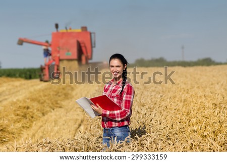 Beautiful girl agronomist with note book standing in golden wheat field and looking at camera, combine harvesting in background