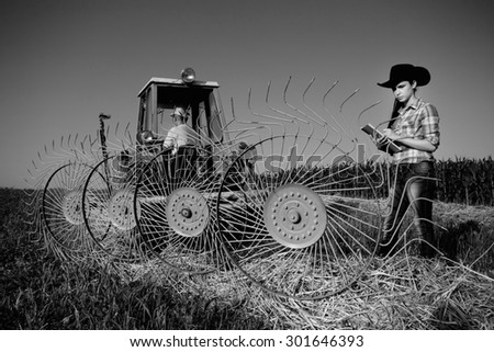Young girl farmer standing beside haying rake attached to tractor, black and white image