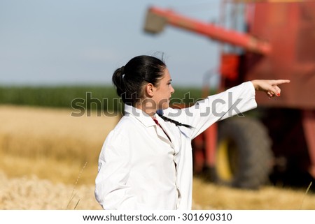 Young woman agronomist standing in golden wheat field in front of combine harvester and showing something with forefinger
