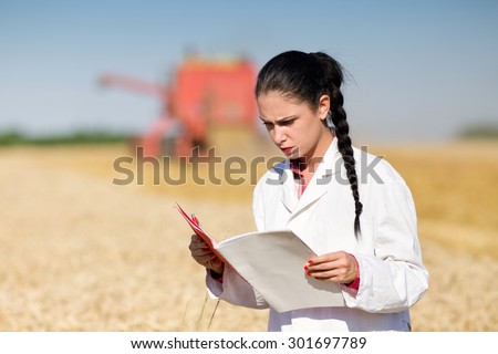Young woman agronomist in white coat reading documentation on wheat field. Combine harvester working in background