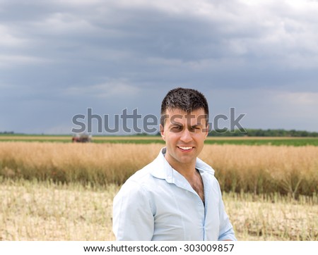 Portrait of young man in shirt walking on rapeseed field during harvest. Tractor in background