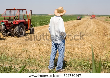 Old man peasant standing on field with combine and tractor in background