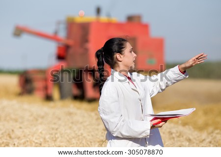Young woman agronomist standing in golden wheat field in front of combine harvester and talking to someone