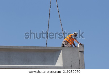 Construction worker standing on concrete beam on height and placing truss lifted by crane