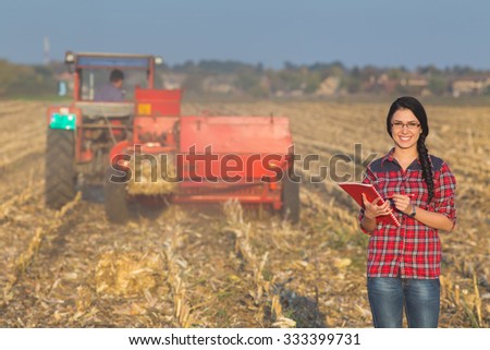 Young woman farmer standing on corn field during baling. Tractor in background