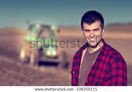Portrait of young handsome farmer on farmland. Tractor working in background