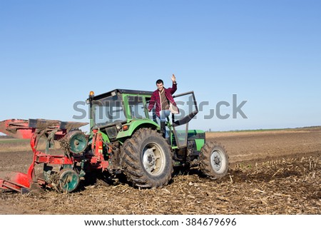 Young farmer standing on tractor in the field and holding arm high in the air
