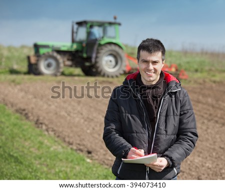Young farmer supervising work and writing notes on farmland, tractor harrowing in background