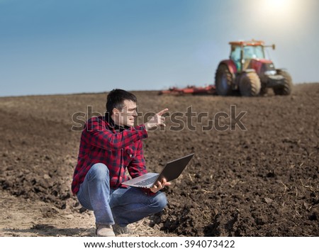 Young farmer with laptop supervising work on farmland, tractor harrowing in background