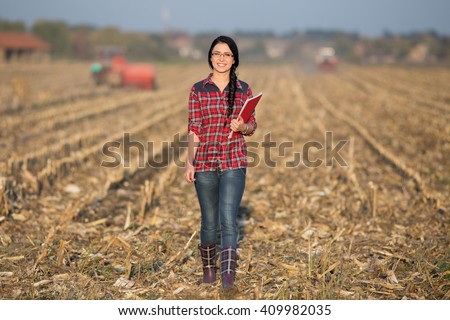 Young woman farmer walking on corn field during baling. Tractor working in background