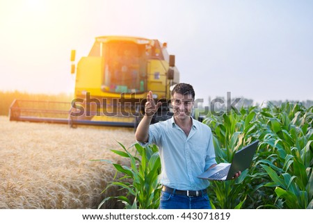 Young attractive farmer holding laptop and standing in corn field while combine harvesting wheat in background
