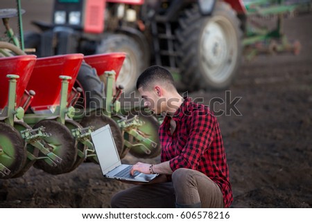 Young farmer with laptop squatting in field in front of tractor with sowing equipment. Smart technology in agriculture