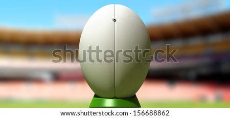 A plain white textured rugby ball on a green kicking tee in a stadium