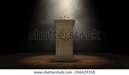 A wooden speech podium with three small microphones attached on a dark background spotlit by a single spotlight