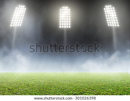 A generic outdoor stadium with an unmarked green grass pitch with an eerie mist at night under illuminated floodlights