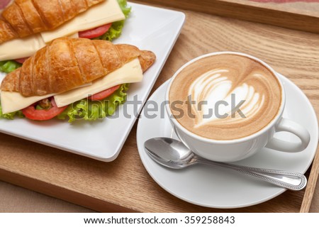A cup of coffee with heart latte art on top and croissant cheese sandwiches
