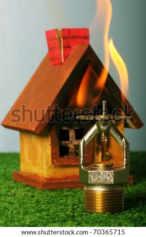 Close up image of fire sprinkler. Fire sprinklers are part of an integrated water piping system designed for life and fire safety. Replica of house on fire added to background.