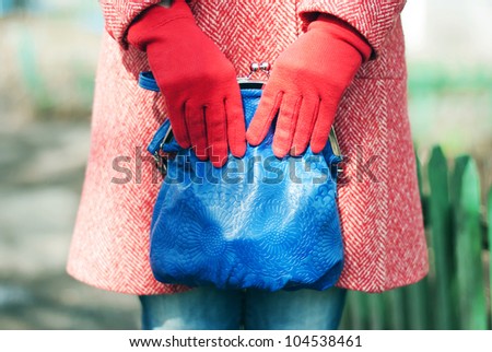 A fashion close up of a woman in a red spring-autumn wool topcoat, blue jeans and red gloves. Woman is holding a blue leather bag in her hands. On the background you can notice a green fence