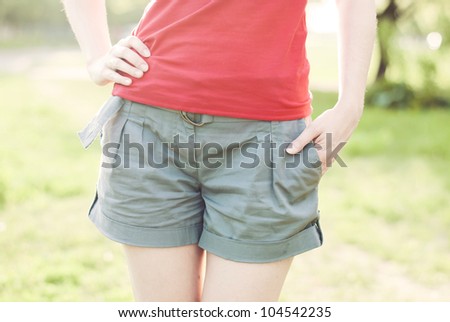 A close up of a woman waist and hands on it on the pastel green summer background in a back light.  Woman is wearing a red t-shirt, grey summer shorts