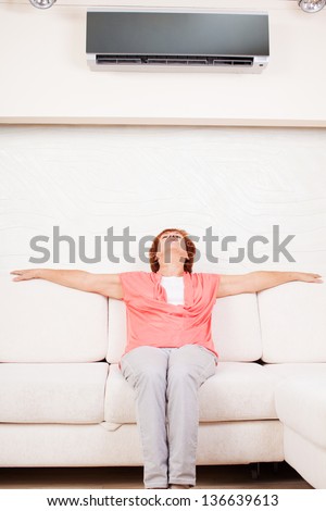 Woman escapes from the heat under the air conditioner at home. Happy mature woman on sofa
