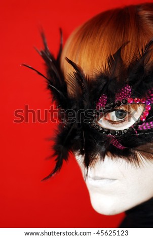 Woman wearing carnival mask,  colored eye lenses and  white makeup looking directly to the camera on red background