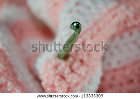 Close-up of the tip of a green crochet needle that is stuck in a pink and white crocheted blanket.  Very narrow depth of field with only the tip of the needle in focus.