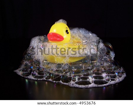 Bright yellow rubber duck bath toy in a small clear glass bowl of water with bubbles and suds pouring over the sides onto the black table. Black background.