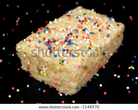 Close up color photograph of a delicious marshmallow crispy dessert bar with colorful sprinkles on and around it.  Black background.