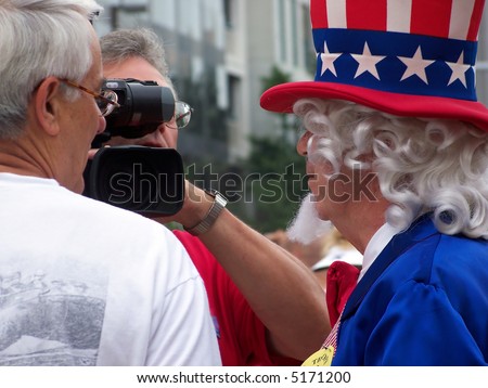 Close up photograph of a man interviewing another man who is dressed in an American patriotic costume.  The third man in the photo is holding a large video camera.