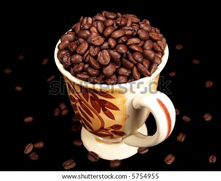 A decorative mug full of rich, dark coffee beans with scattered beans on the table around the it.  Black background.