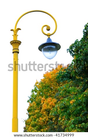 Blue street lamp on a yellow lamppost hanging over the trees against the white sky