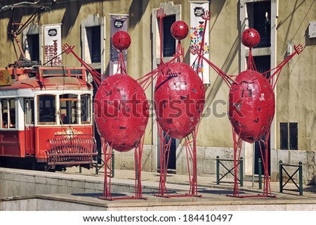 LISBON, PORTUGAL - JULY 24, 2013: Traditional red tram and three red people statues in Placa do Municipio. The authorship of the abstract sculpture is unknown.