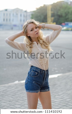 Blond girl in a  blue jeans shorts and white blouse  posing on the street