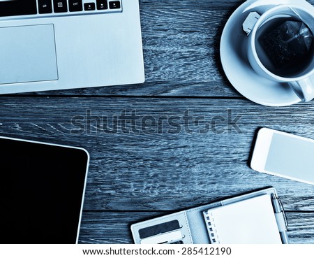 Office desk with laptop computer, tablet pc, planner, pen, mobile smartphone and cup of tea.  Blue toned