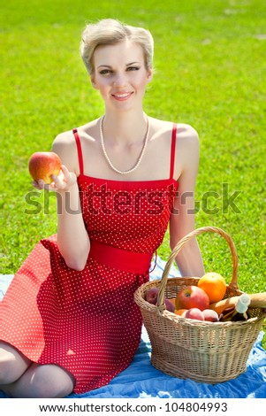 Young blond woman is sitting on grass. Basket with bread, fruit and bottle of vine is standing near her