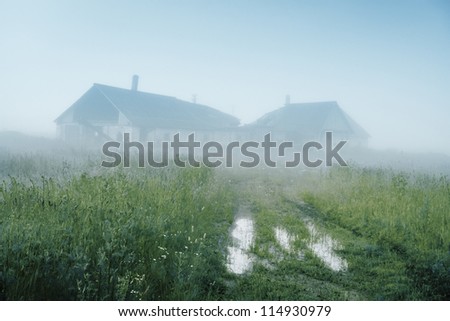 Picturesque farm ruins on hill in fog in morning. Road with puddles in deserted area in morning with fog