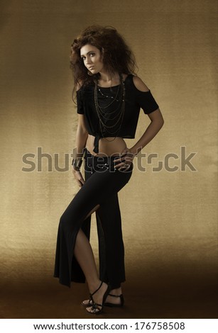 Beautiful woman with wild auburn hair posing in a revealing black outfit with gladiator sandals against a golden background