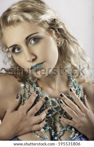 Portrait of blonde woman wearing blue makeup and nail polish, holding her hands up to her elaborate beaded blue necklace