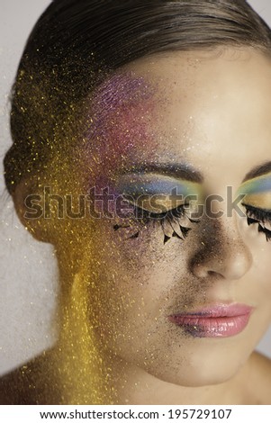 Portrait of beautiful woman wearing colorful makeup, long false eyelashes, pink lipstick, sitting with her eyes closed while pink, purple and golden glitter is thrown against the side of her face