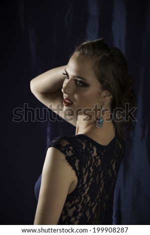 Sensual brunette woman wearing navy lace dress looking back over her shoulder against a dark navy drape