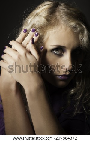 Blonde woman with striking blue eyes gazing daringly from behind her hands with purple nail polish on the nails.