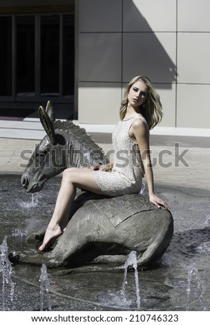 Lovely blonde woman posing on bronze water feature of urban fountain in the afternoon sunlight looking glamorous and beautiful,