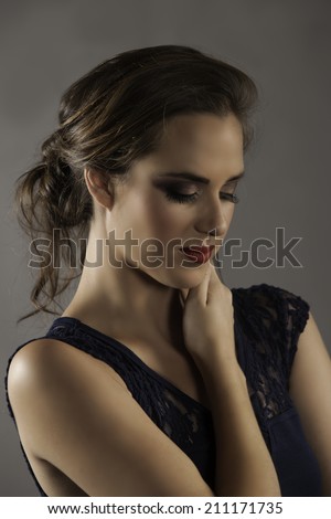 Portrait of beautiful brunette woman holding her hand to her neck, wearing lace navy dress and dramatic makeup with her long brown hair in a messy bun