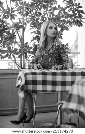 Monochrome image of sexy blonde woman wearing black stilettos and leopard print blouse, seated outside at a restaurant table with her black handbag resting on it