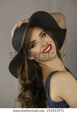 Portrait of gorgeous young woman wearing red lipstick, navy dress and hat, posing with her hair in a loose, curly style while smiling happily