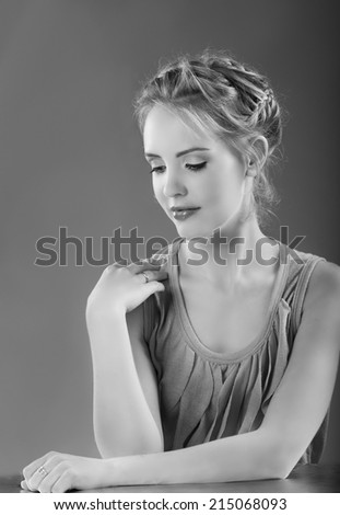 Monochrome portrait in black and white of lovely blonde woman with braided hair, natural makeup and a feminine top looking down while touching her shoulder